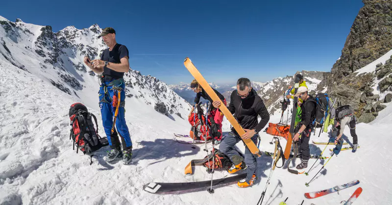 Vier mensen maken zich klaar voor een skitocht op een besneeuwde berghelling; ze stellen hun uitrusting en ski’s af onder een strakblauwe hemel, met ruige bergtoppen op de achtergrond.